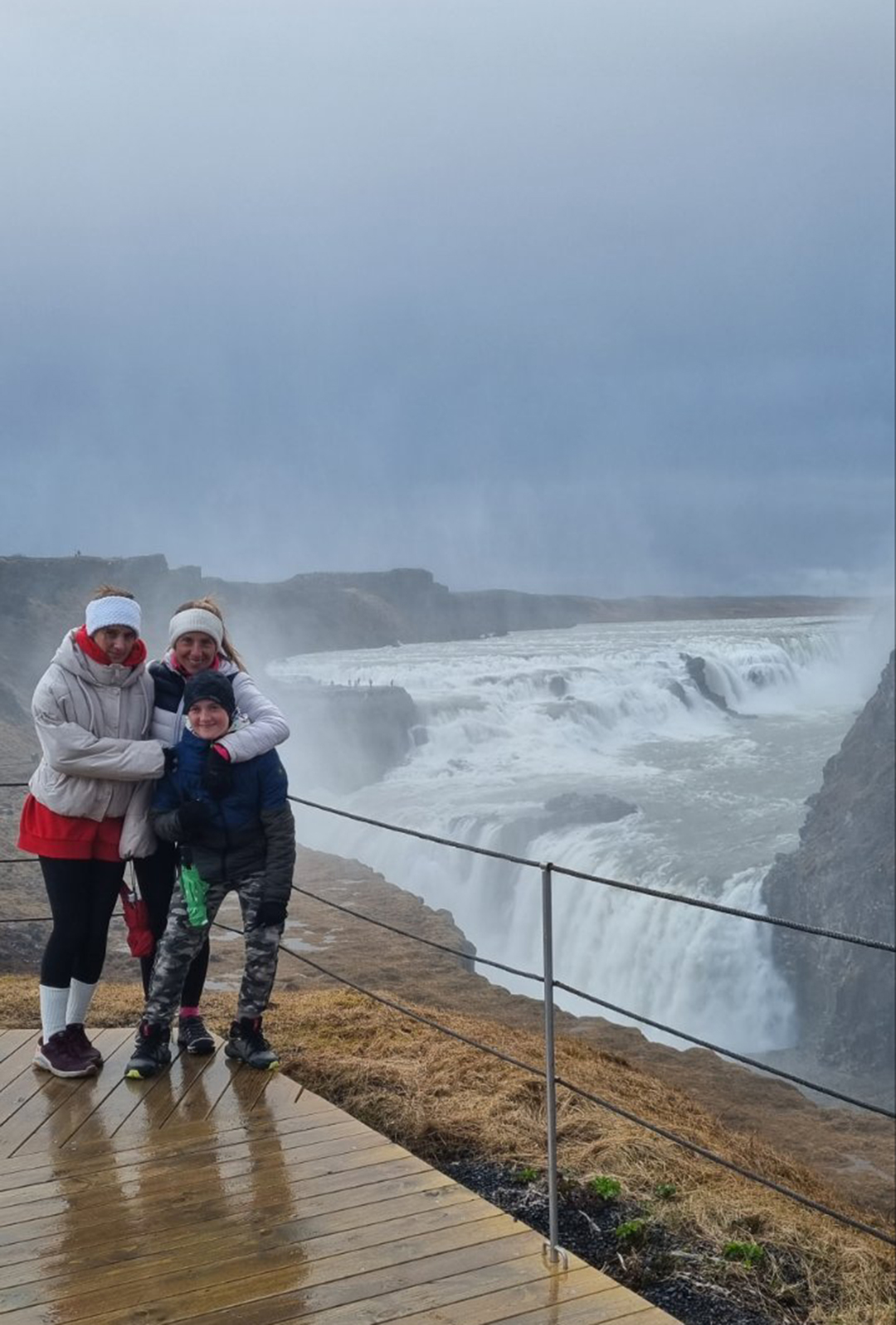 A family standing at a viewpoint overlooking the powerful Gullfoss waterfall, surrounded by mist, wind and dramatic scenery.