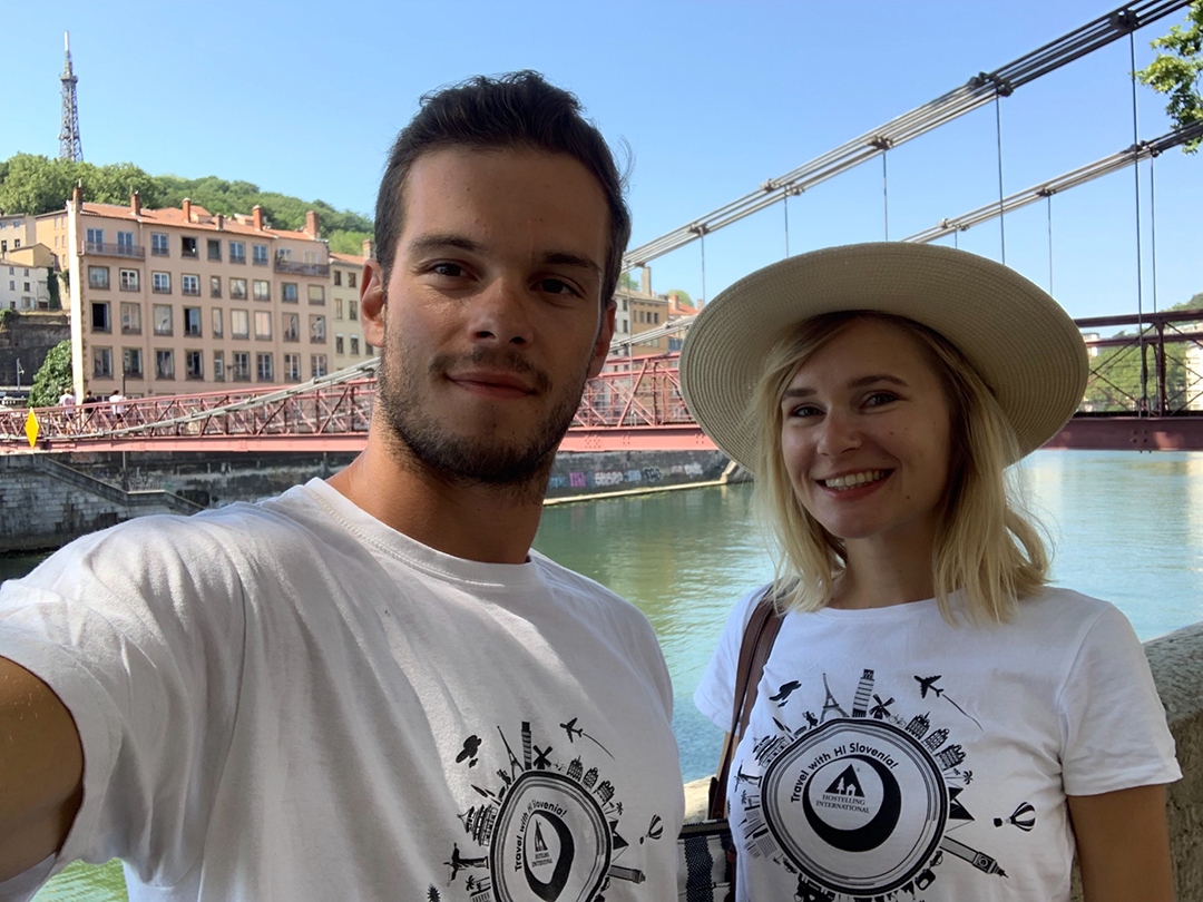 Two volunteers wearing Hostelling International Slovenia T-shirts posing by the river, representing a sense of community and belonging.
