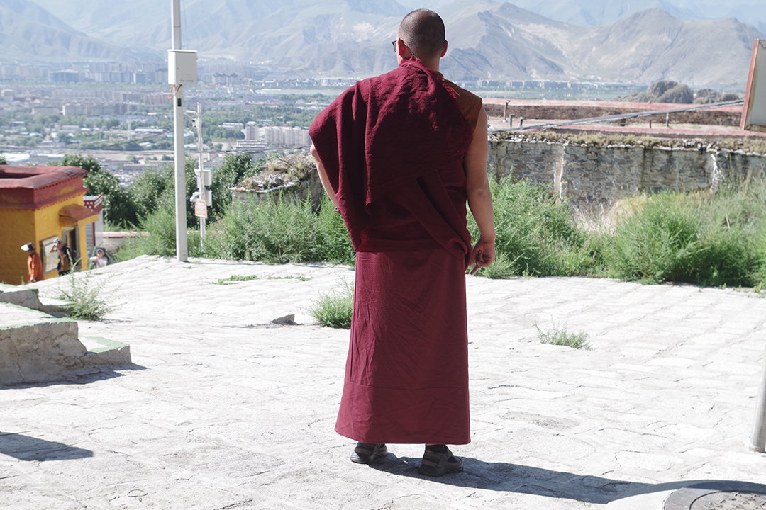 Budistični menih v tradicionalnem rdečem oblačilu s pogledom na Lhaso in gorato tibetansko pokrajino.