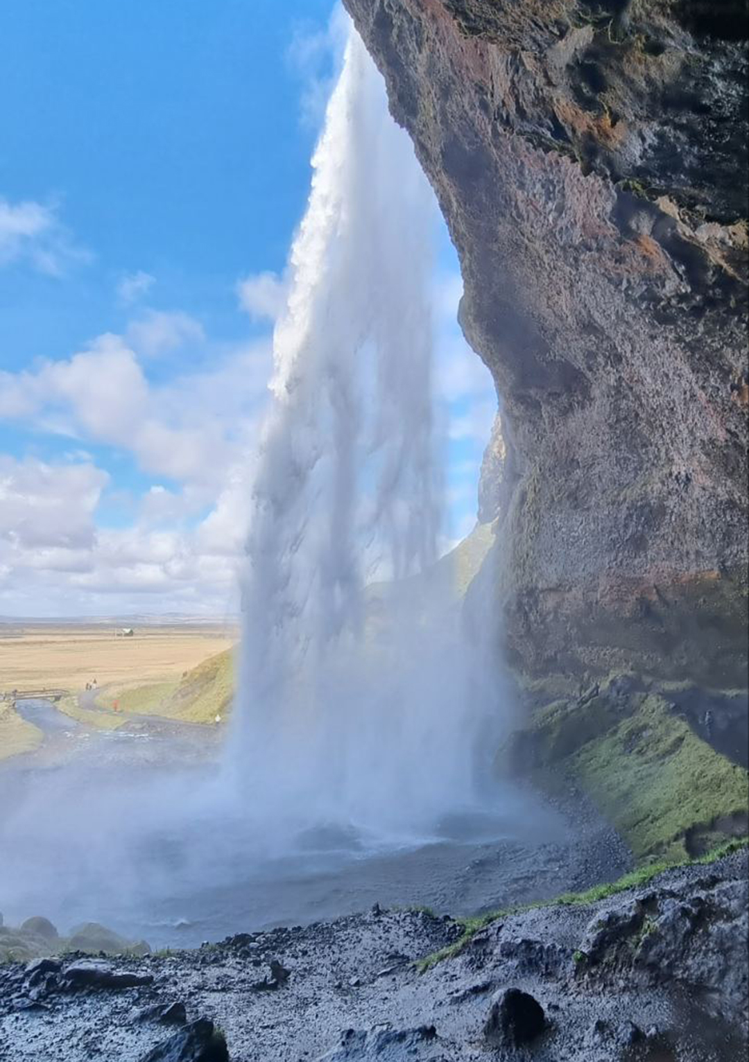 Slap Seljalandsfoss na Islandiji, kjer pot vodi za padajočo vodo, s pogledom na odprto islandsko pokrajino in mogočno naravo juga otoka.